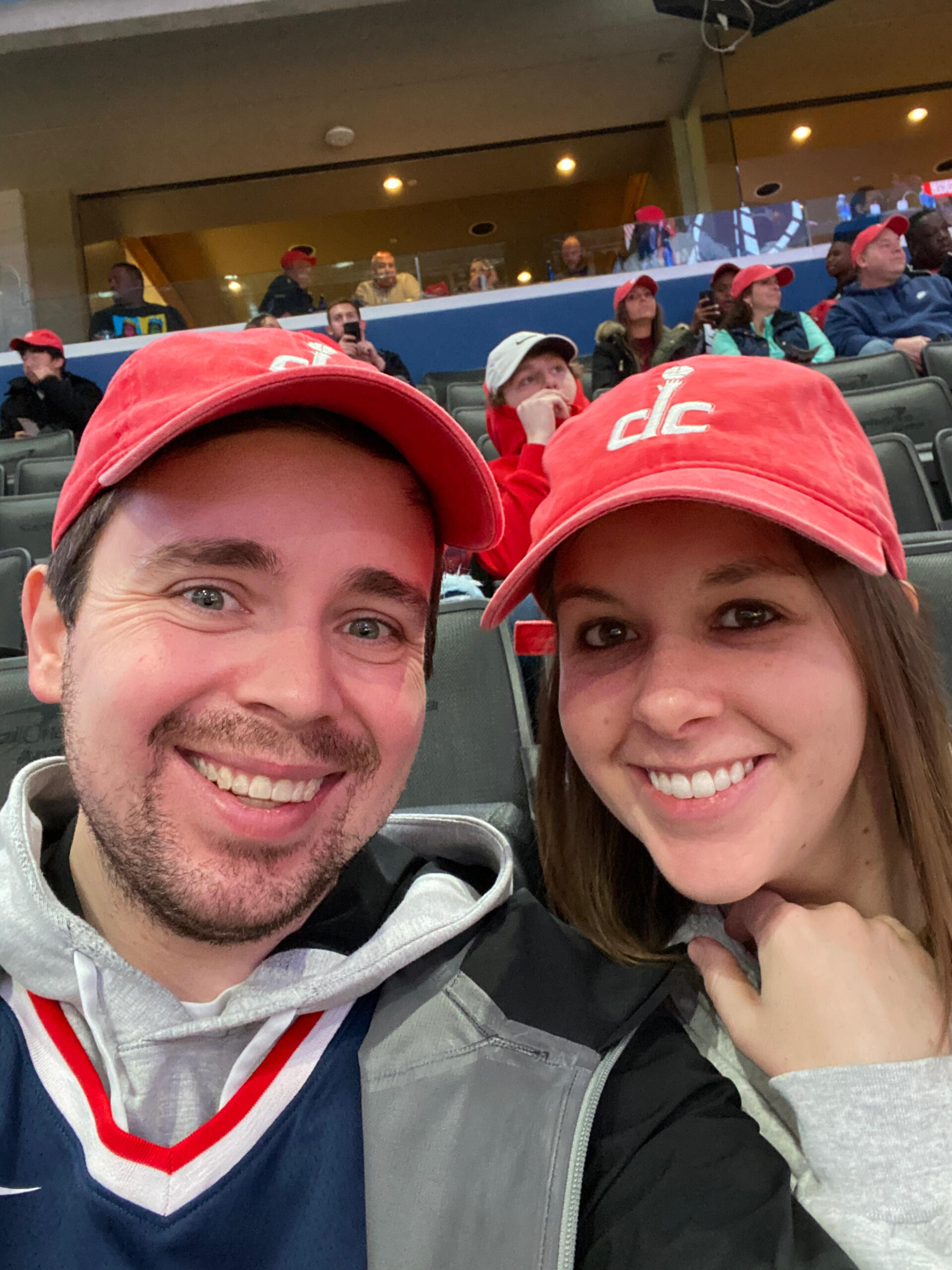 Ethan Wechtaluk smiling with his wife Courtney at a basketball game, both wearing red Washington Wizards caps and sitting in the stands with fans in the background.