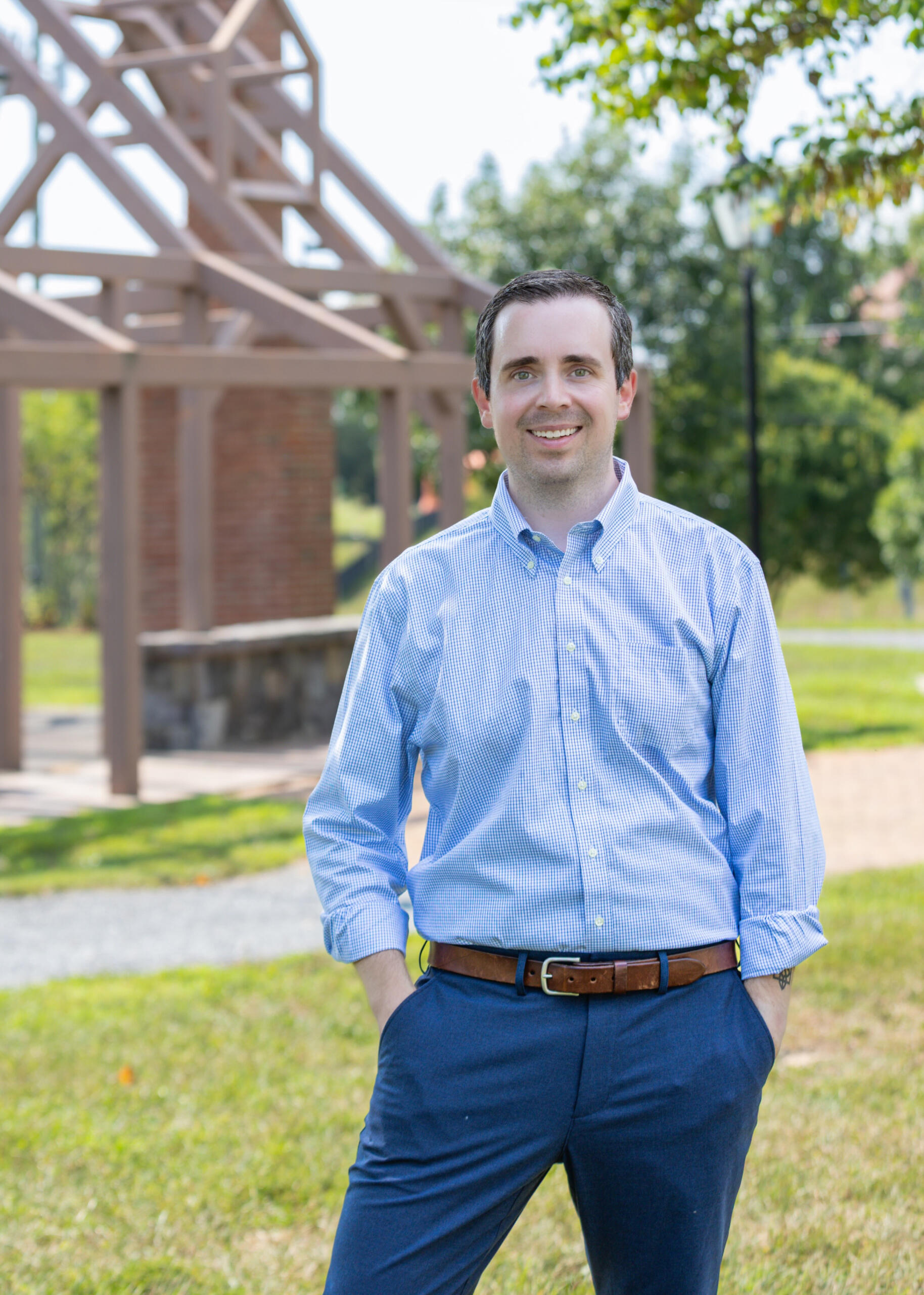 Ethan Wechtaluk standing outdoors in front of a wooden and brick pavilion structure, wearing a light blue button-down shirt and navy pants, smiling with hands in pockets on a sunny day.